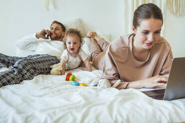 Frau benutzt Laptop, während der Mann mit seiner Tochter auf dem Bett zu Hause telefoniert - VPIF04295