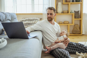 Geschäftsmann arbeitet am Laptop, während seine Tochter auf dem Sofa im Wohnzimmer auf dem Schoß schläft - VPIF04318