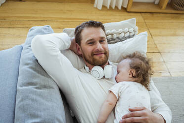 Lächelnder Mann mit Hand hinter dem Kopf, der sich entspannt, während seine Tochter zu Hause schläft - VPIF04325