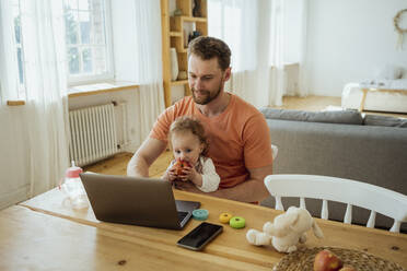 Man working on laptop while daughter eating apple on lap at home - VPIF04340