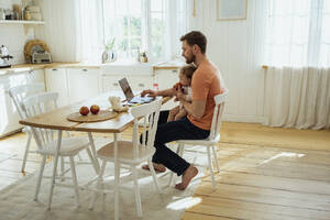 Businessman feeding daughter while working on laptop at home - VPIF04341