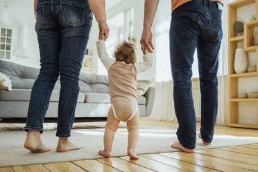 Baby girl holding hands of parents while walking in living room - VPIF04346