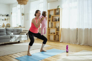 Smiling woman carrying daughter while exercising in living room at home - VPIF04355