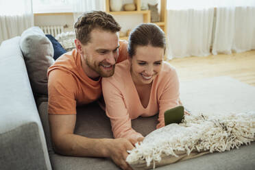 Lächelndes Paar, das ein Mobiltelefon benutzt, während es zu Hause auf dem Sofa liegt - VPIF04360