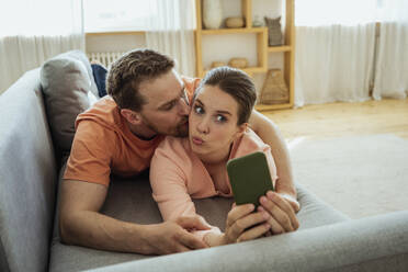 Man kissing woman taking selfie through mobile phone while lying on sofa at home - VPIF04361