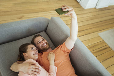 Lächelnder Mann, der ein Selfie mit einer Frau über sein Smartphone macht, während er zu Hause auf dem Sofa liegt - VPIF04362
