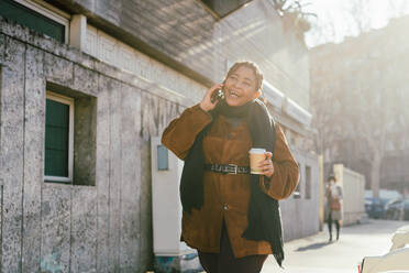 Italy, Smiling woman talking on phone and holding disposable cup on city street - ISF24699