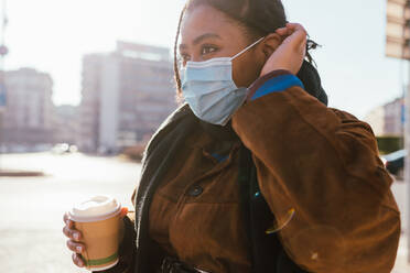 Italy, Young woman in face mask holding disposable cup outdoors - ISF24700