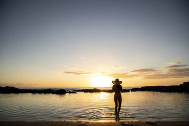 Eine Frau steht in der Untiefe und trägt einen Hut bei Sonnenuntergang - CAVF94395