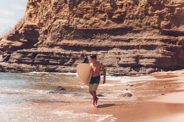 Boy walking on the beach carrying his surfboard - cliffs on the back. - CAVF94407