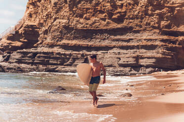 Junge, der am Strand spazieren geht und sein Surfbrett trägt - Klippen auf dem Rücken. - CAVF94407