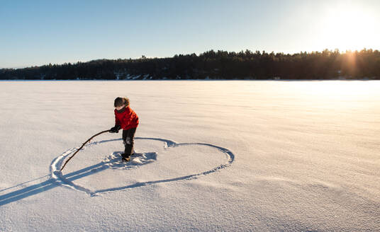 Ein Kind zeichnet ein Herz auf einem verschneiten Feld in der Morgensonne. - CAVF94421
