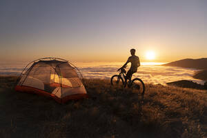 Mountainbiker steht bei Sonnenuntergang neben dem Zelt und genießt die Aussicht - CAVF94515