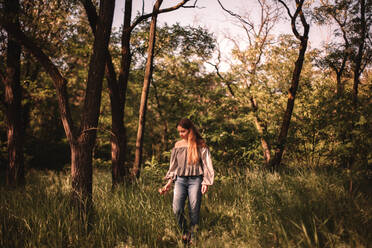 Teenage girl walking through green grass in forest during summer - CAVF94521