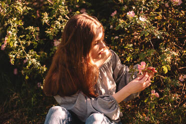 Teenage girl holding flower sitting in summer park during sunny day - CAVF94526