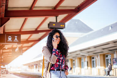 Happy young African girl talking on cell phone at train station in Europe - CAVF94561