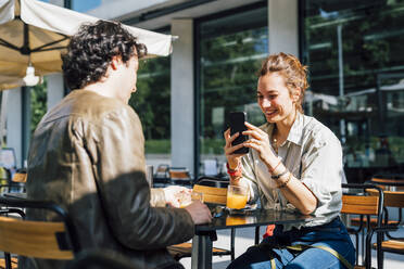 Smiling woman taking photo of boyfriend through smart phone at sidewalk cafe - MEUF03184