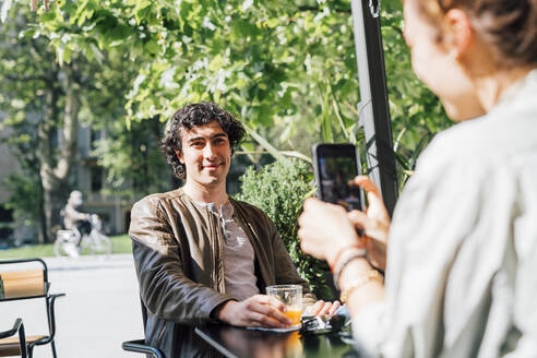 Freundin fotografiert ihren Freund mit dem Handy in einem Straßencafé - MEUF03185