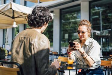 Frau mit Sonnenbrille und Mobiltelefon, während sie mit einem Mann in einem Straßencafé sitzt - MEUF03188