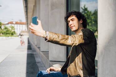 Junger Mann, der ein Selfie mit seinem Smartphone macht, während er im Freien sitzt - MEUF03198