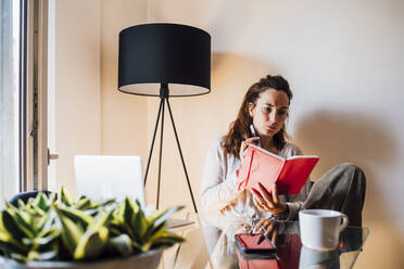 Woman holding book while sitting by dining table at home - MEUF03220