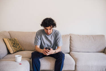 Mann, der ein Mobiltelefon benutzt, während er mit einer Kaffeetasse auf dem Sofa sitzt - MEUF03222
