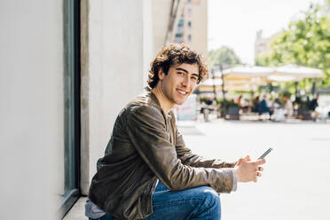 Smiling man holding mobile phone while sitting at doorway - MEUF03242