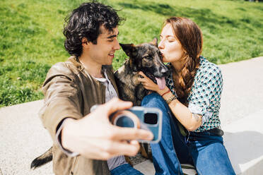 Mann nimmt Selfie durch Smartphone mit Frau küssen Hund während sonnigen Tag - MEUF03254