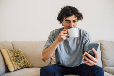 Man drinking coffee while using mobile phone at home - MEUF03274