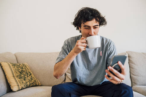 Mann trinkt Kaffee, während er zu Hause sein Mobiltelefon benutzt - MEUF03274