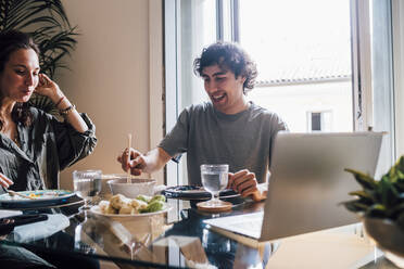 Smiling man eating food while sitting by woman at home - MEUF03287