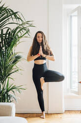 Woman practicing yoga while standing on one leg at home - MEUF03299