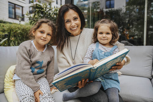 Lächelnde Mutter mit süßer Tochter, die ein Buch auf dem Sofa im Hinterhof hält - MFF08230