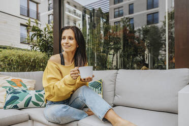 Smiling woman with mug while looking away on sofa at backyard - MFF08239