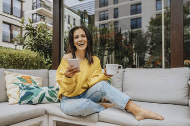 Woman laughing while holding mug and smart phone on sofa at backyard - MFF08240