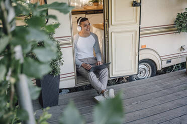 Smiling male freelancer sitting with laptop at camper doorway - VPIF04395