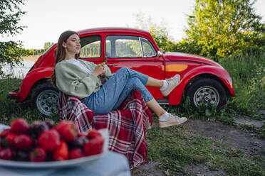 Young woman looking away while sitting on chair by red car - VPIF04398