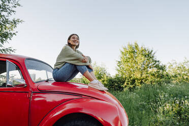 Lächelnde junge Frau, die auf der Motorhaube eines Autos sitzt und die Knie umarmt - VPIF04400