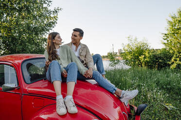 Lächelndes junges Paar, das sich auf der Motorhaube eines roten Autos sitzend ansieht - VPIF04402