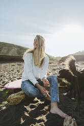 Smiling blond woman looking away while sitting by dog at beach - MTBF01049