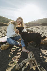 Junge blonde Frau, die ihren Hund streichelt, während sie auf einem Baumstamm am Strand sitzt - MTBF01050