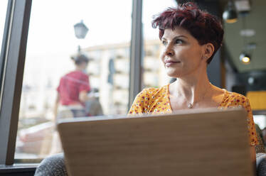 Ältere Frau mit Laptop schaut durch das Fenster eines Cafés - JCCMF03109