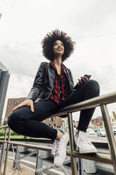 Smiling woman with mobile phone sitting on railing - MEUF03308