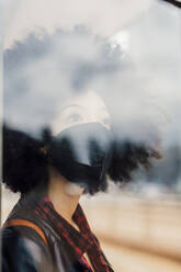 Thoughtful woman with protective face mask looking through glass wall - MEUF03313