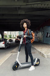 Young woman standing with electric push scooter on road under bridge - MEUF03319
