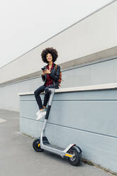 Young woman with mobile phone sitting on retaining wall by electric push scooter - MEUF03324