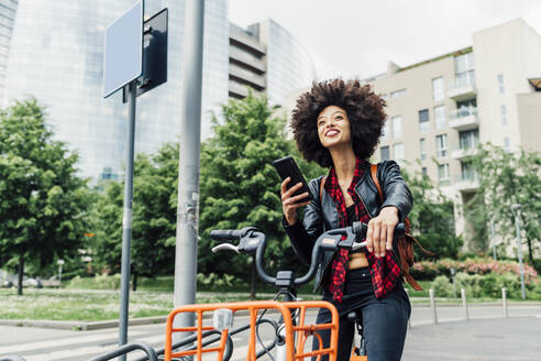 Junge Frau entriegelt ihr Fahrrad mit dem Smartphone an einer Parkstation - MEUF03329