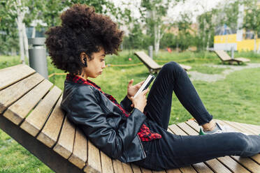 Woman using mobile phone while sitting on wooden chair at park - MEUF03360