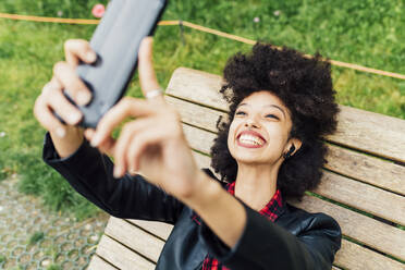 Glückliche Frau, die ein Selfie mit ihrem Mobiltelefon macht, während sie auf einem Stuhl im Park liegt - MEUF03365