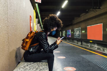 Frau mit Gesichtsschutz, die in einer U-Bahn-Station sitzt und ein Mobiltelefon benutzt - MEUF03379